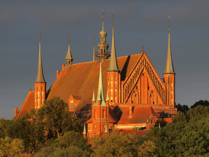 Bazylika archikatedralna we Fromborku widziana z oddala przy zachodzącym słońcu. Olbrzymi, ceramiczny dach warmińskiej hali z narożnymi wieżyczkami wbudowanymi w korpus świątyni kontrastuje do zachmurzonego nieba i zadrzewienia Wzgórza Katedralnego. (Sierpień 2011)